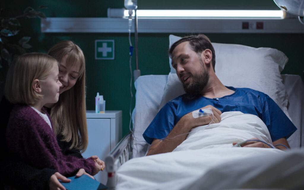 A man in a hospital bed being visited by his wife and daughter