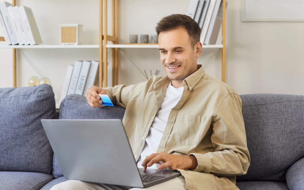 Man sat on a grey sofa with a laptop and card managing his savings