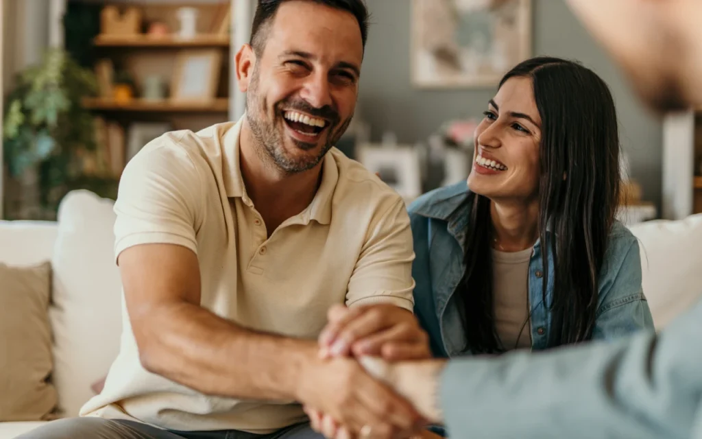 Man and woman sitting on a sofa smiling. the man is reaching out his hand to shake hands warmly withy another person