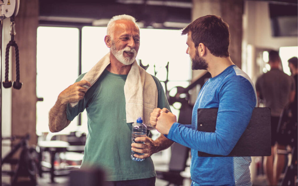 A man with a grey beard, water bottle and towel talking to a coach in a gym