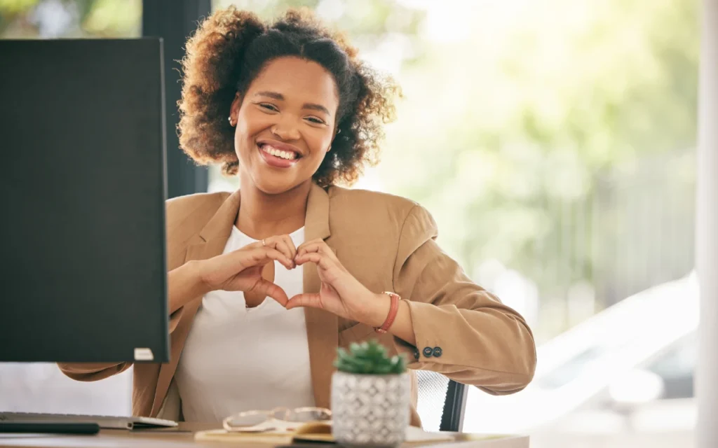 Woman sat at a computer smiling giving a sign of a heart with both hands